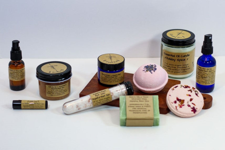 A collection of products on a white background, including salt scrub, serum, lip balm, body butter, soap, bath bomb, and candle