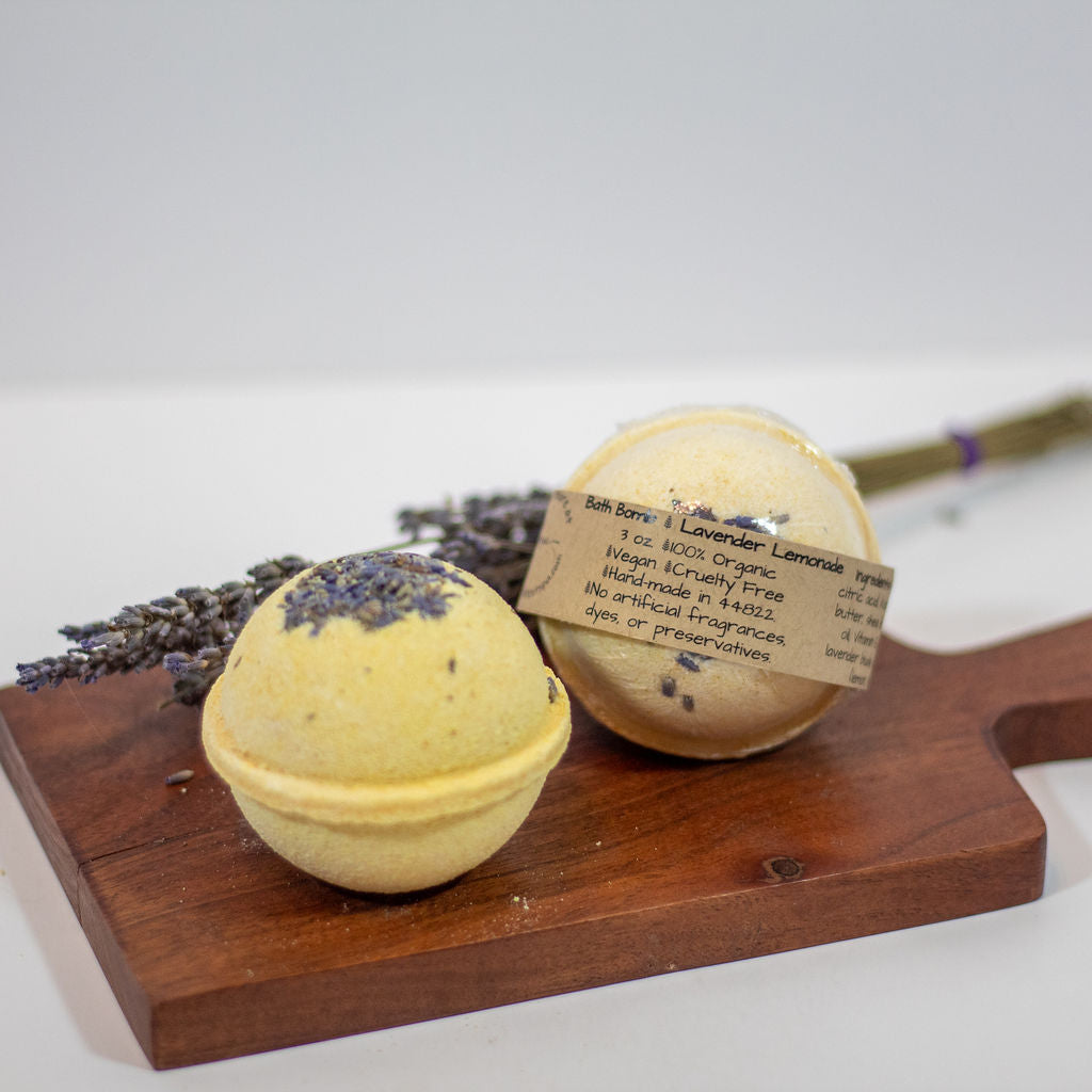 Two round bath bombs on a wooden board, sprinkled with dried lavender one wrapped in a paper band labeled 'Lavender Lemonade'.