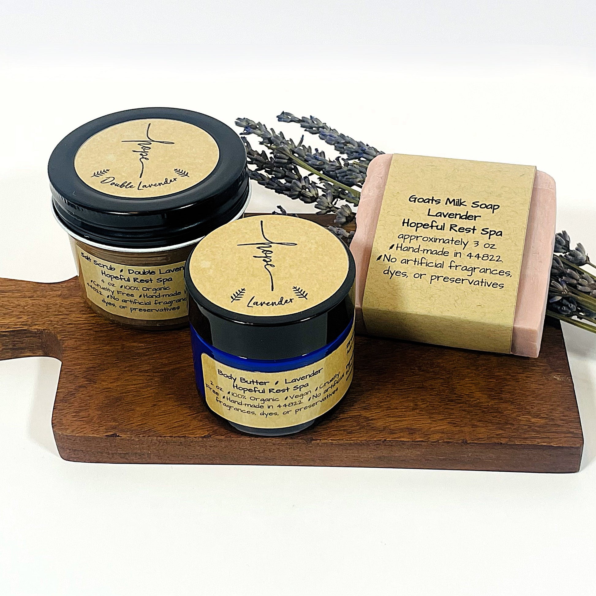 A gift set including a jar of lavender goats milk bar soap, a pink salt scrub, and a jar of 2 oz. lavender body butter, all with labels and placed on a wooden tray with a bundle of lavender.