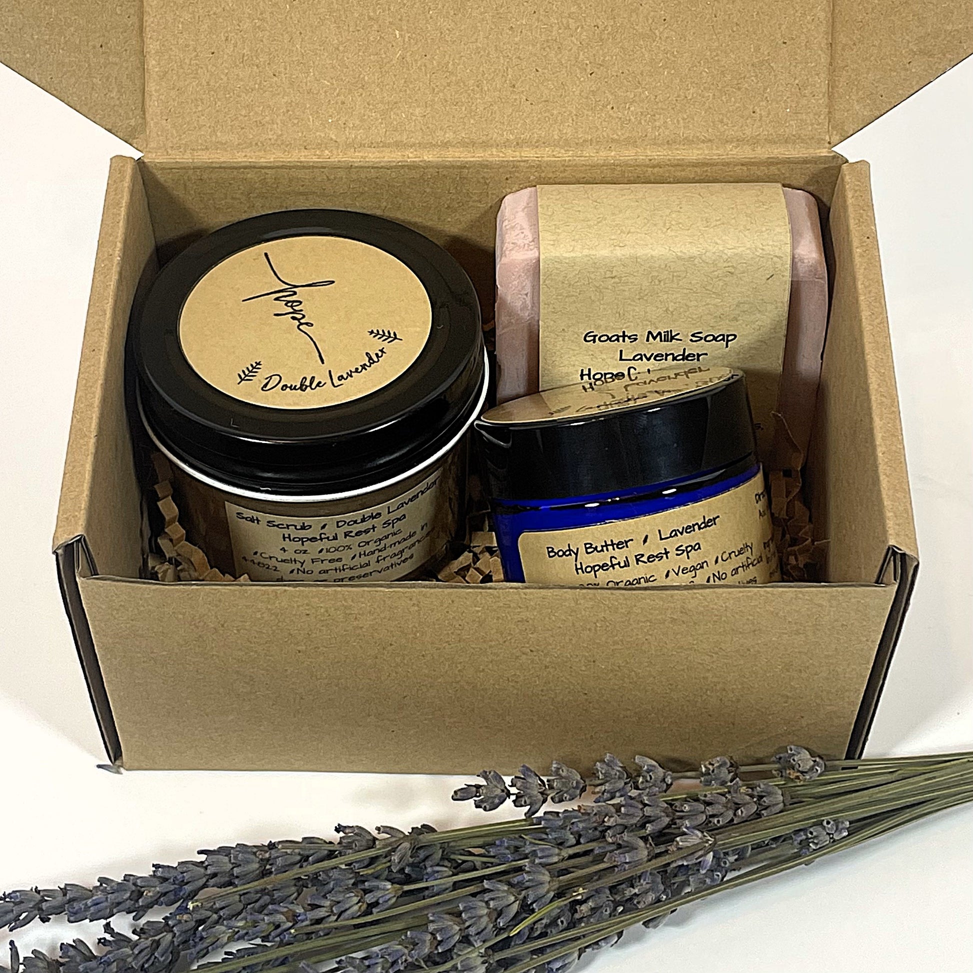 A gift set including a jar of lavender goats milk bar soap, a pink salt scrub, and a jar of 2 oz. lavender body butter, all with labels in a box with a bundle of lavender.