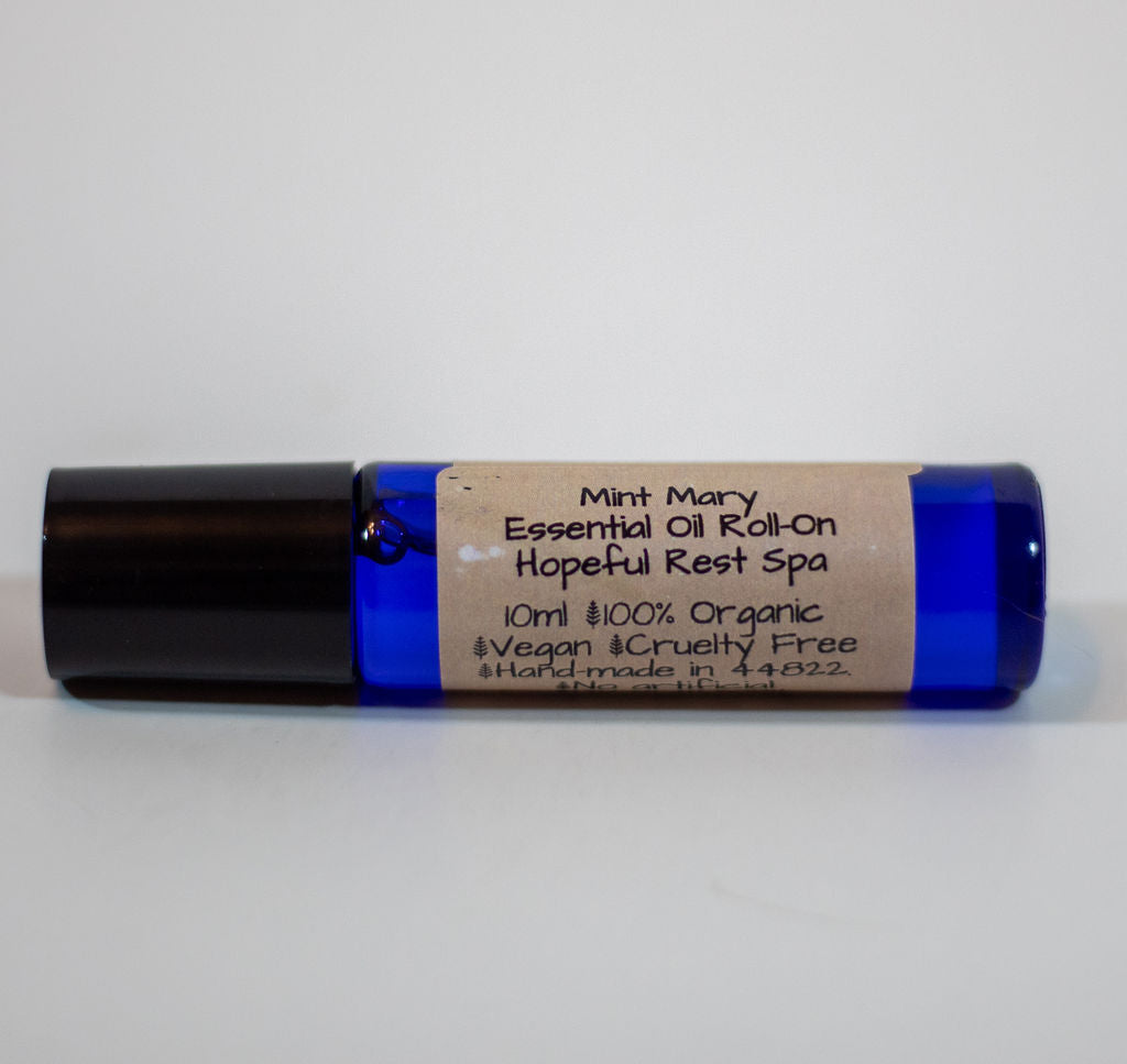 A 10ml essential oil roll-on bottle with a cobalt blue glass body, a black cap, and a brown kraft paper label that reads 'Mint Mary Essential Oil Roll-On Hopeful Rest Spa'.