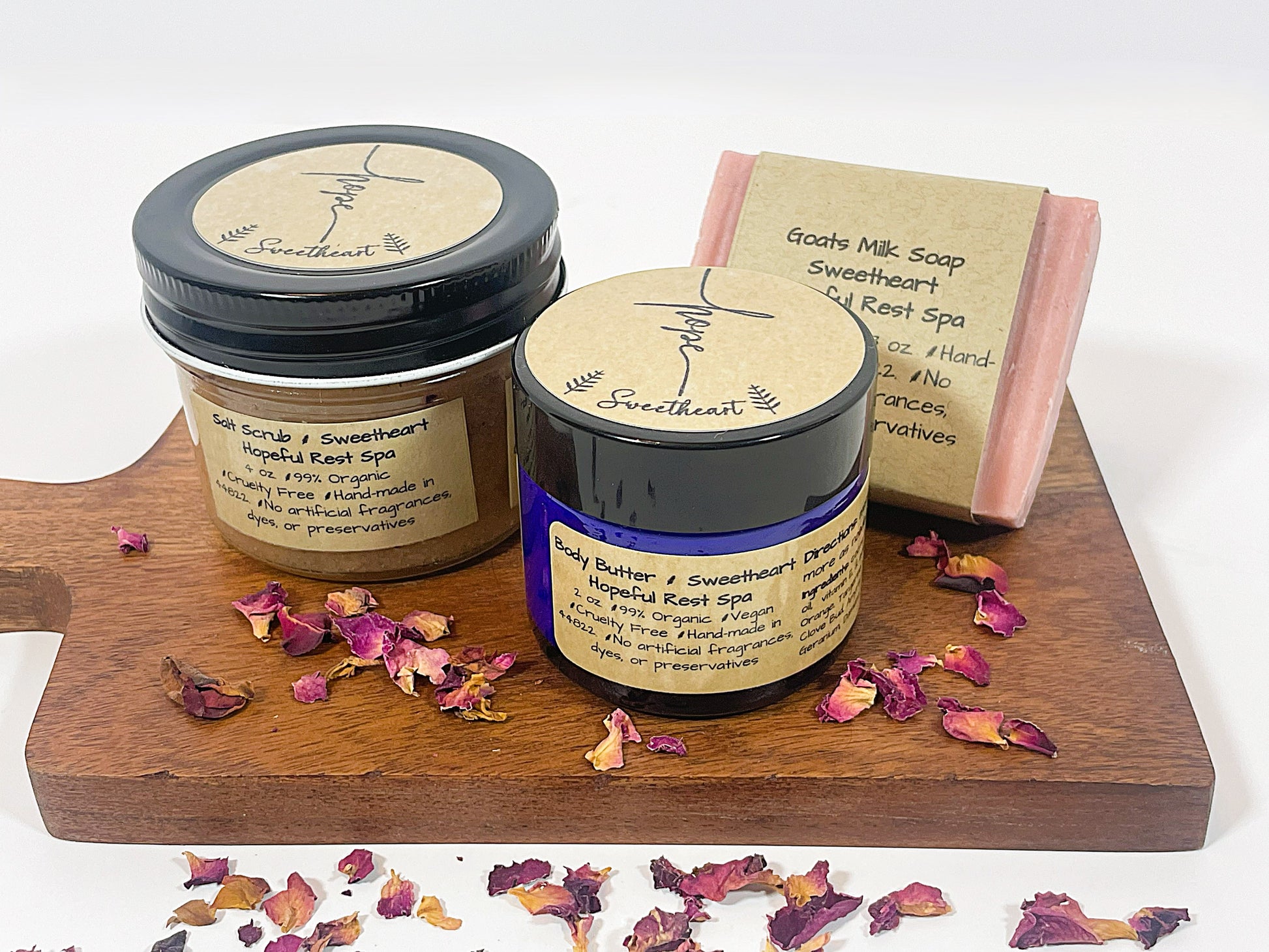 A gift set including a jar of Sweetheart Goats Milk Bar Soap, a jar of Sweetheart Salt Scrub, and a bar of Sweetheart Body Butter, all with labels and placed on a wooden board with petals scattered around.