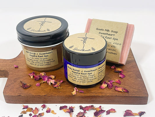 A gift set including a jar of Sweetheart Goats Milk Bar Soap, a jar of Sweetheart Salt Scrub, and a bar of Sweetheart Body Butter, all with labels and placed on a wooden board with petals scattered around.