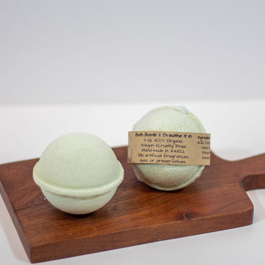 Two pale mint green bath bombs on a wooden board, with one showing a kraft paper label that reads 'Bath Bomb Breathe It in'.