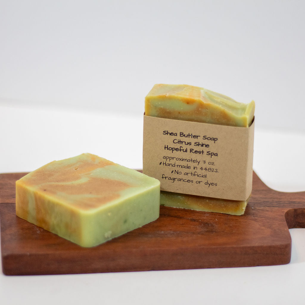 Two handmade soap bars with kraft paper sleeves are displayed on a wooden cutting board against a white background. The soap has a marbled green and orange-yellow texture, and the label reads 'Shea Butter Soap Citrus Shine Hopeful Reef Spa'.