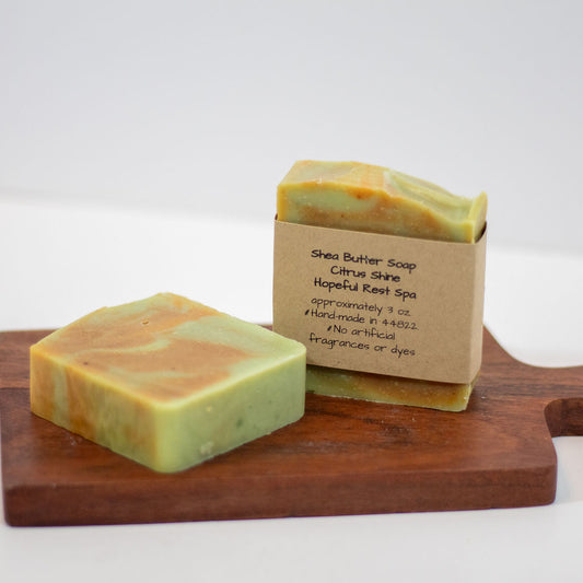 Two handmade soap bars with kraft paper sleeves are displayed on a wooden cutting board against a white background. The soap has a marbled green and orange-yellow texture, and the label reads 'Shea Butter Soap Citrus Shine Hopeful Reef Spa'.
