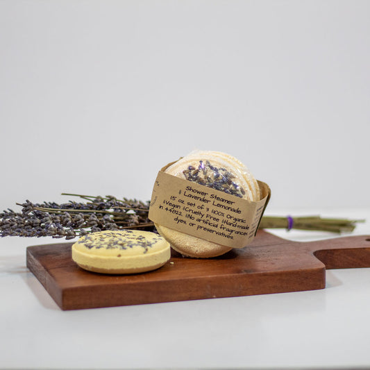 A handmade shower steamer set displayed on a wooden cutting board with dried lavender. The set is wrapped in a kraft paper label that reads 'Lavender Lemonade'.