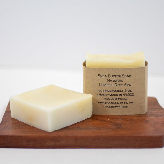 Two rectangular bars of Shea Butter Soap on a wooden board, with one bar in front and one behind in a kraft paper sleeve. The background is a clean white studio backdrop.